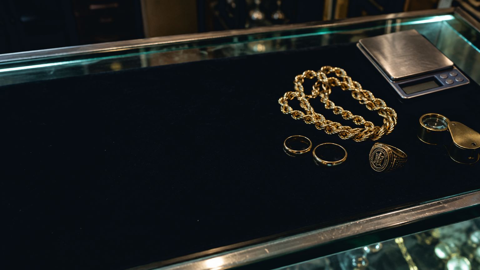 A pawn shop's glass counter after closing: a gold rope chain, two wedding bands and a class ring on black velvet, a jewelry scale and a brass loupe.